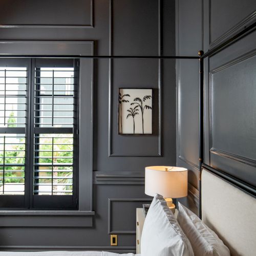 A stylish bedroom with dark walls, a window with shutters, white bedding, a bedside lamp, and a small framed picture of palm trees.