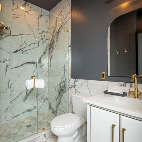 A modern bathroom with a marble shower, gold fixtures, a toilet, and a white vanity with a sink and mirror.