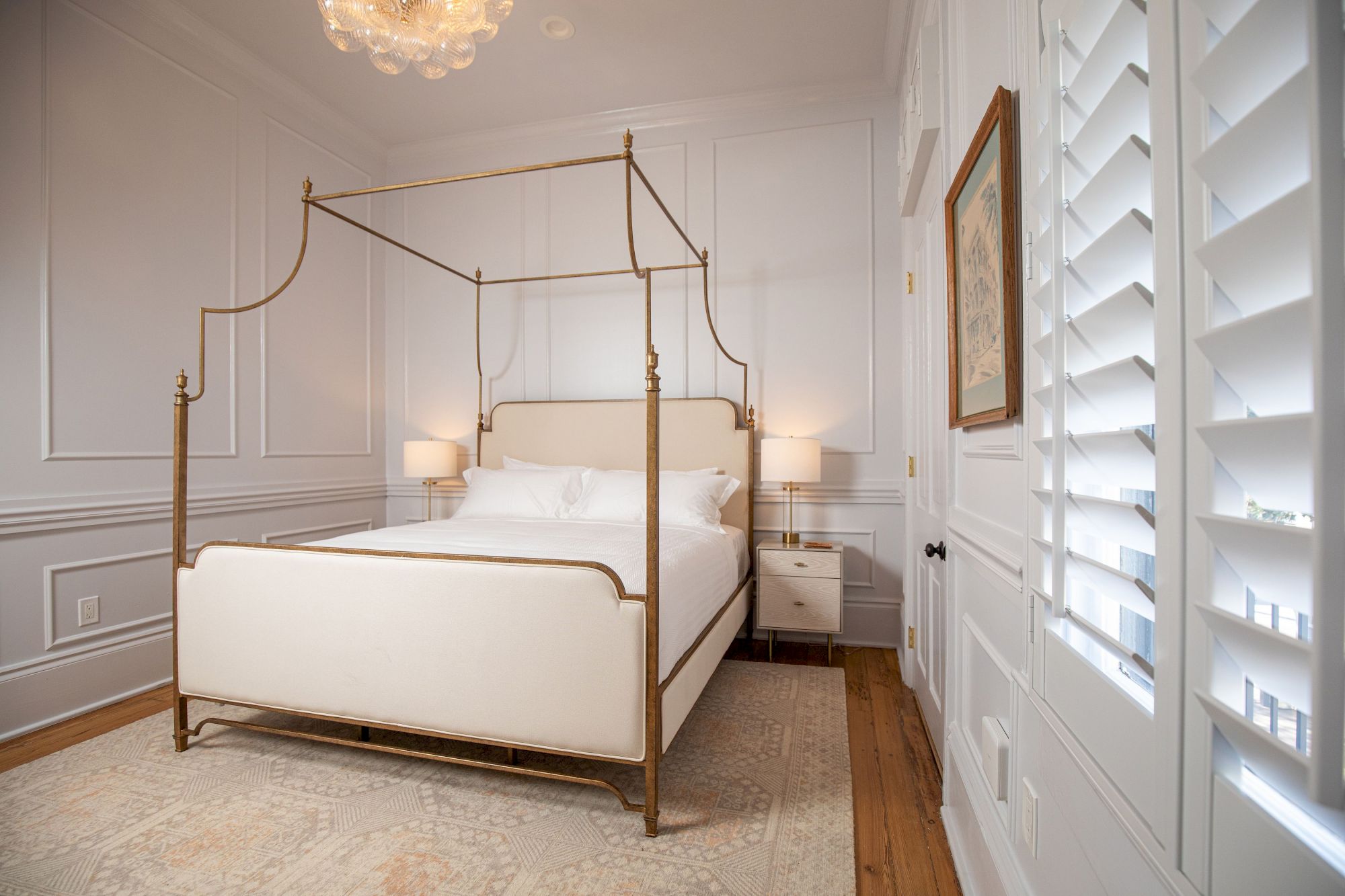 A bedroom with a canopy bed, two side tables, lamps, paneled walls, a rug, and a window with shutters on the right wall.