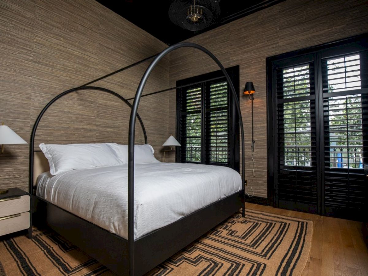 A bedroom with a canopy bed, white linens, side tables, lamps, striped rug, and large windows with shutters.