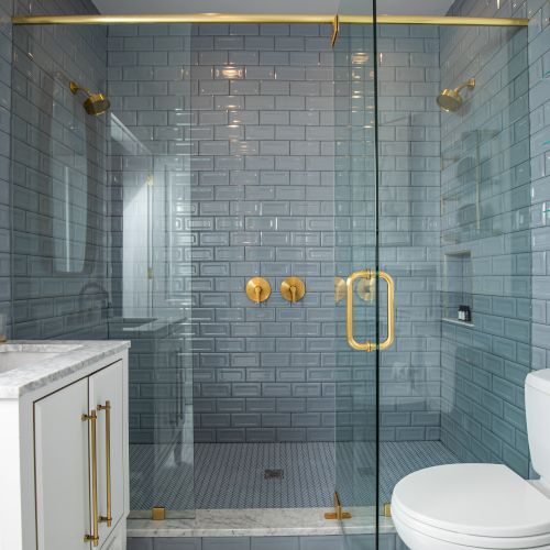 A modern bathroom with a glass shower, gold fixtures, white vanity, and a toilet, featuring gray subway tiles and towel racks.