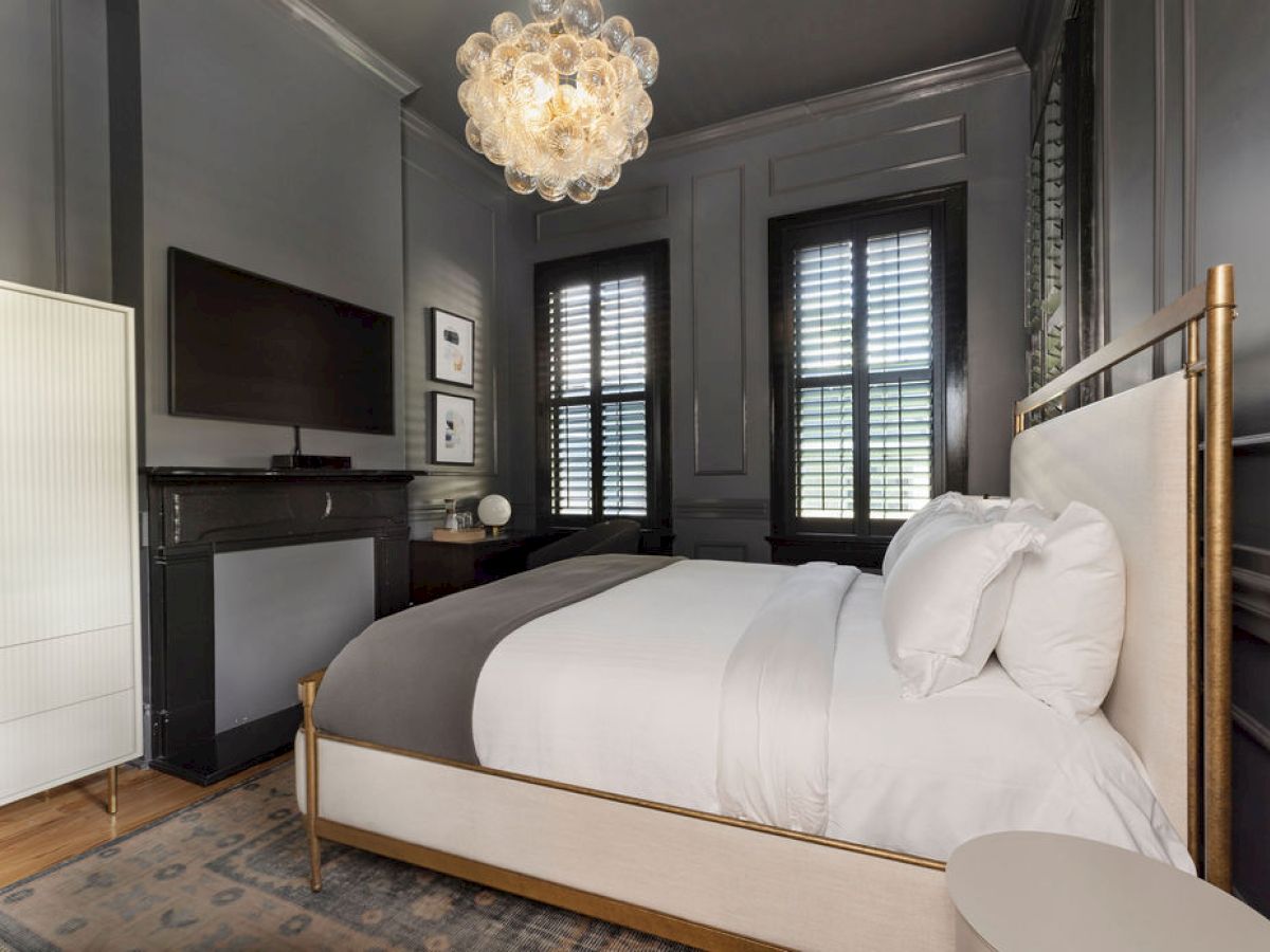 A modern bedroom featuring a bed, elegant chandelier, dark walls, artwork, and a wardrobe with light streaming through window shutters.