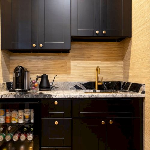A small kitchenette with black cabinets, a marble countertop, a coffee machine, and a mini fridge stocked with beverages ends the scene.