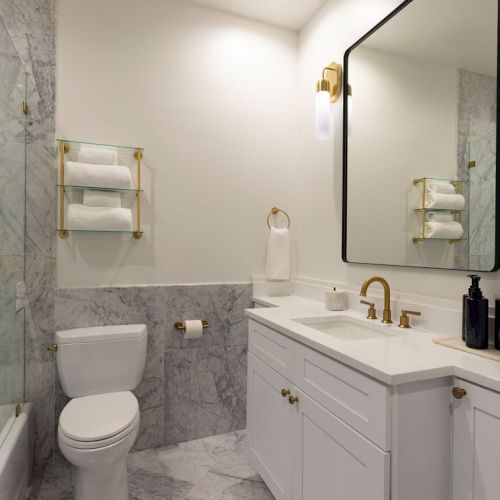 A modern bathroom with a toilet, marble surfaces, white vanity, gold fixtures, large mirror, and glass shower enclosure.