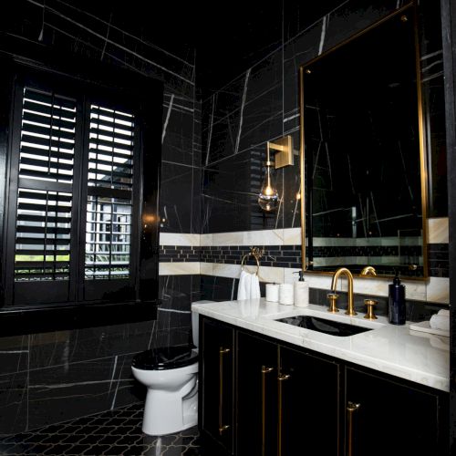 A modern bathroom with dark walls, a white countertop, a large mirror, and a toilet. The design features gold accents and unique lighting.