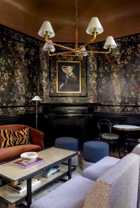 Cozy room with floral wallpaper, a framed photo, chandelier, various seating, round tables, and a coffee table with books.
