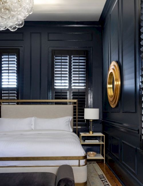 A bedroom with dark walls, a bed, chandelier, lamp, and a round wall decor. It features shuttered windows and a nightstand.