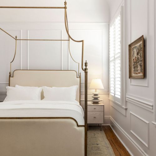 A stylish bedroom with a four-poster bed, white linens, a chandelier, and a nightstand with a lamp against paneled walls.