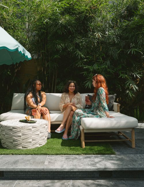 Three people are sitting on a patio sofa, surrounded by lush greenery, engaging in conversation in a relaxed outdoor setting.