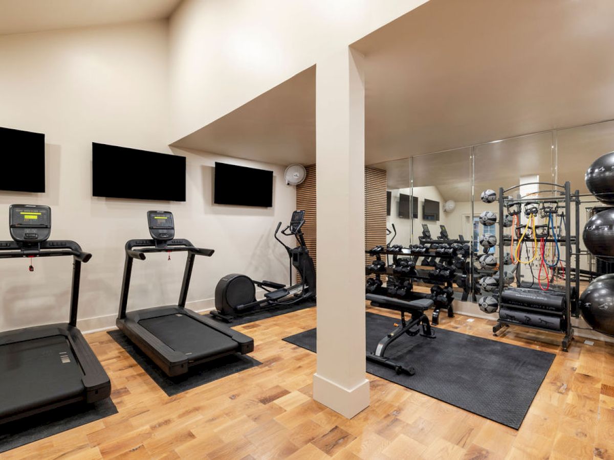 The image shows a home gym with treadmills, an elliptical, weights, exercise balls, and mirrors on a wooden floor.