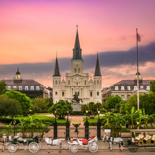 A historic cathedral stands amid lush greenery and a colorful sky, with horse-drawn carriages lined up in front of a decorative iron fence.