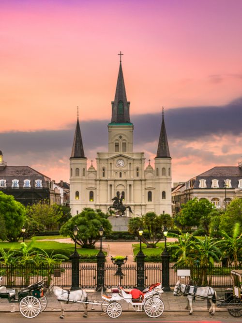 A historic cathedral stands amid lush greenery and a colorful sky, with horse-drawn carriages lined up in front of a decorative iron fence.