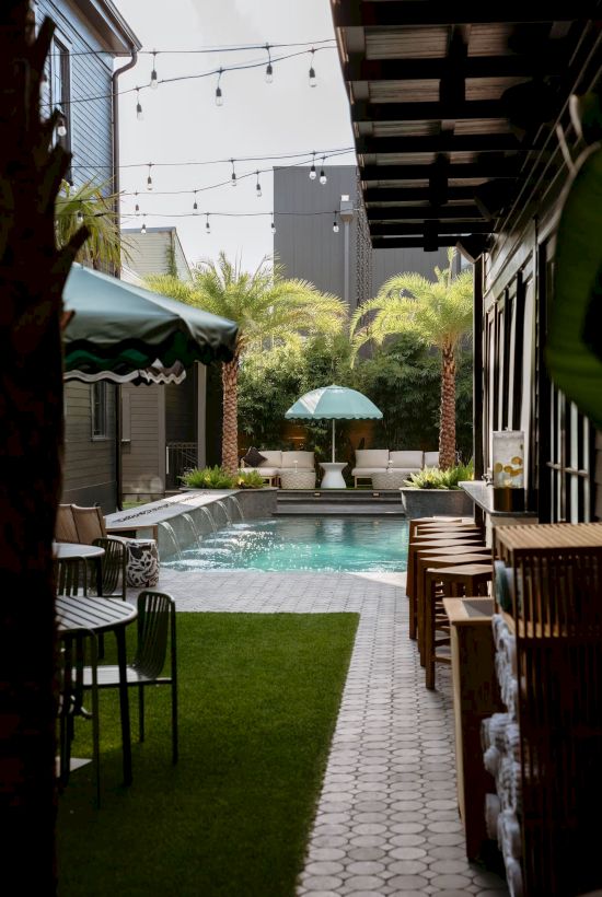 A modern outdoor pool area with palm trees, string lights, and a walkway. Tables and chairs are set up for relaxation and dining.