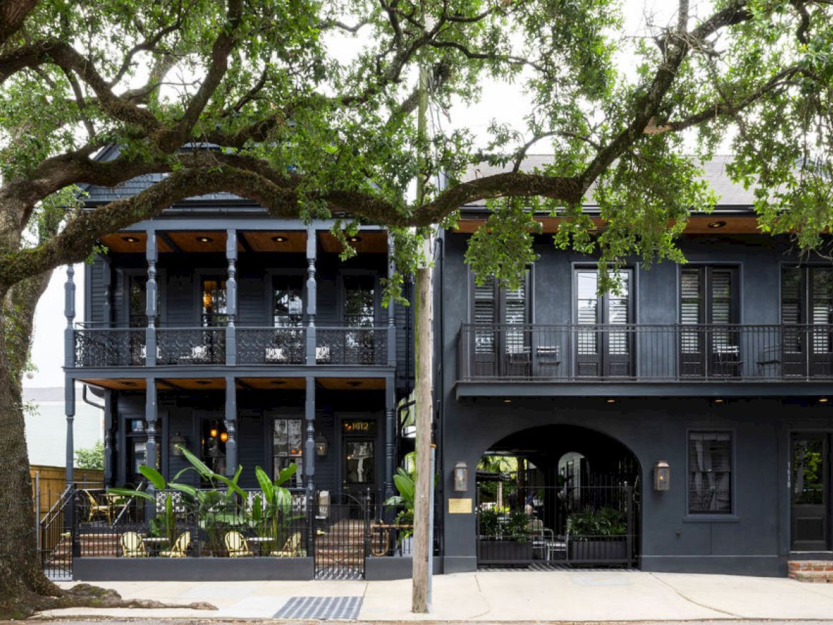 The image shows a two-story, dark-colored building with balconies, surrounded by trees and plants, featuring an archway on the ground floor.
