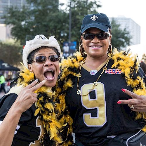Two people wearing sports jerseys and feather boas pose energetically outdoors, with buildings and trees in the background.